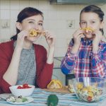 Joyful mother and cute cheerful daughter have fun grimacing silly with vegetables and looking into camera while cooking in the kitchen at home. Family, cook, and people concept